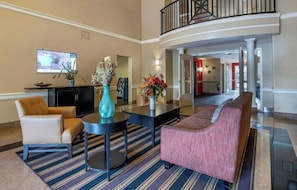 A lobby with a sofa, armchair, and a small table with a vase. There is a television mounted on the wall. - Studio 6 Suites Jacksonville Deerwood Park FL (Jacksonville)
