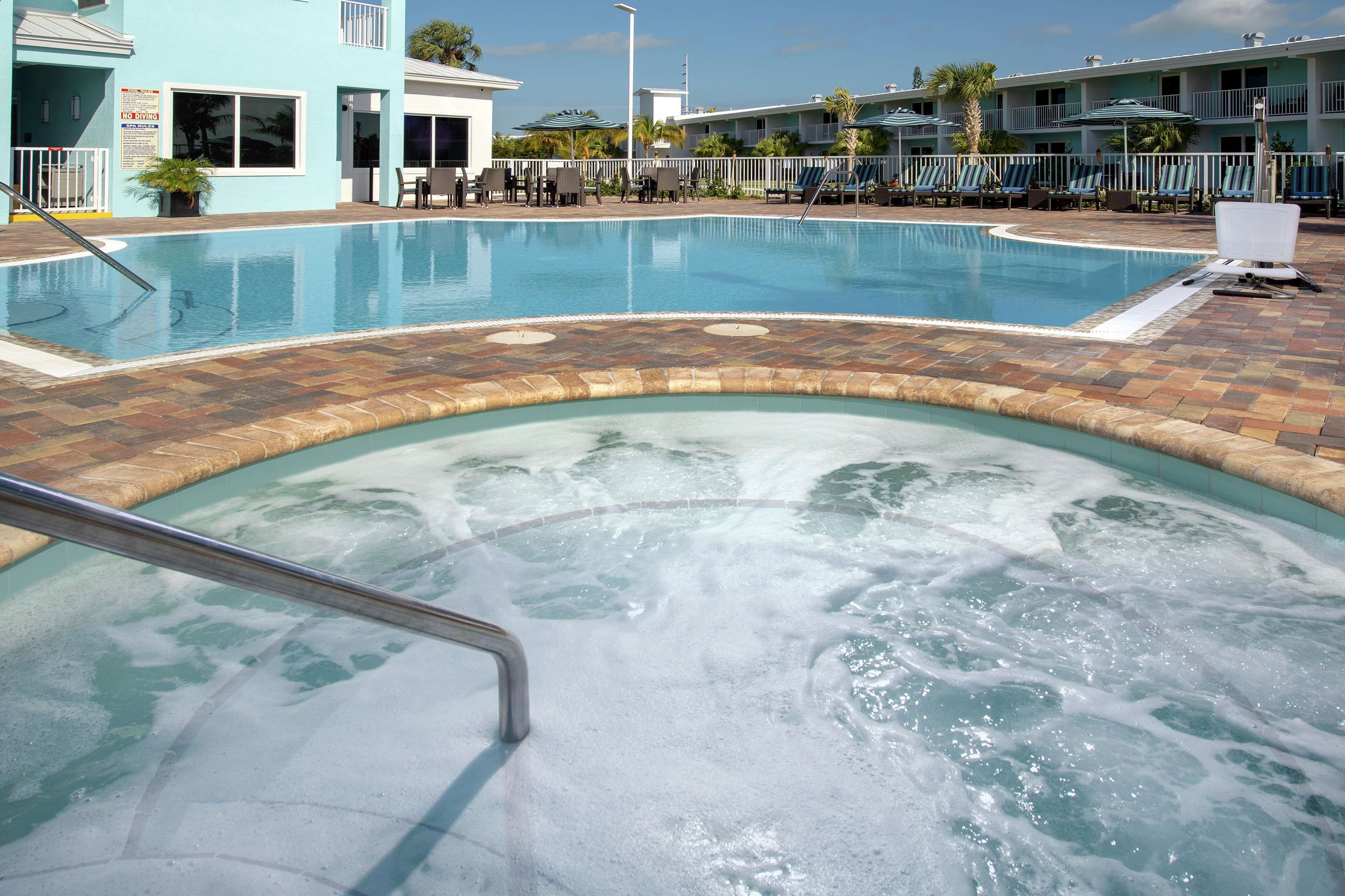 Een zwembad met een jacuzzi, ligbedden en een gebouw met een balkon op de achtergrond.