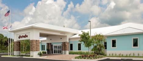 A Hampton Inn hotel with a modern design, featuring a welcoming entrance, a parking lot, and a clear sky with clouds.