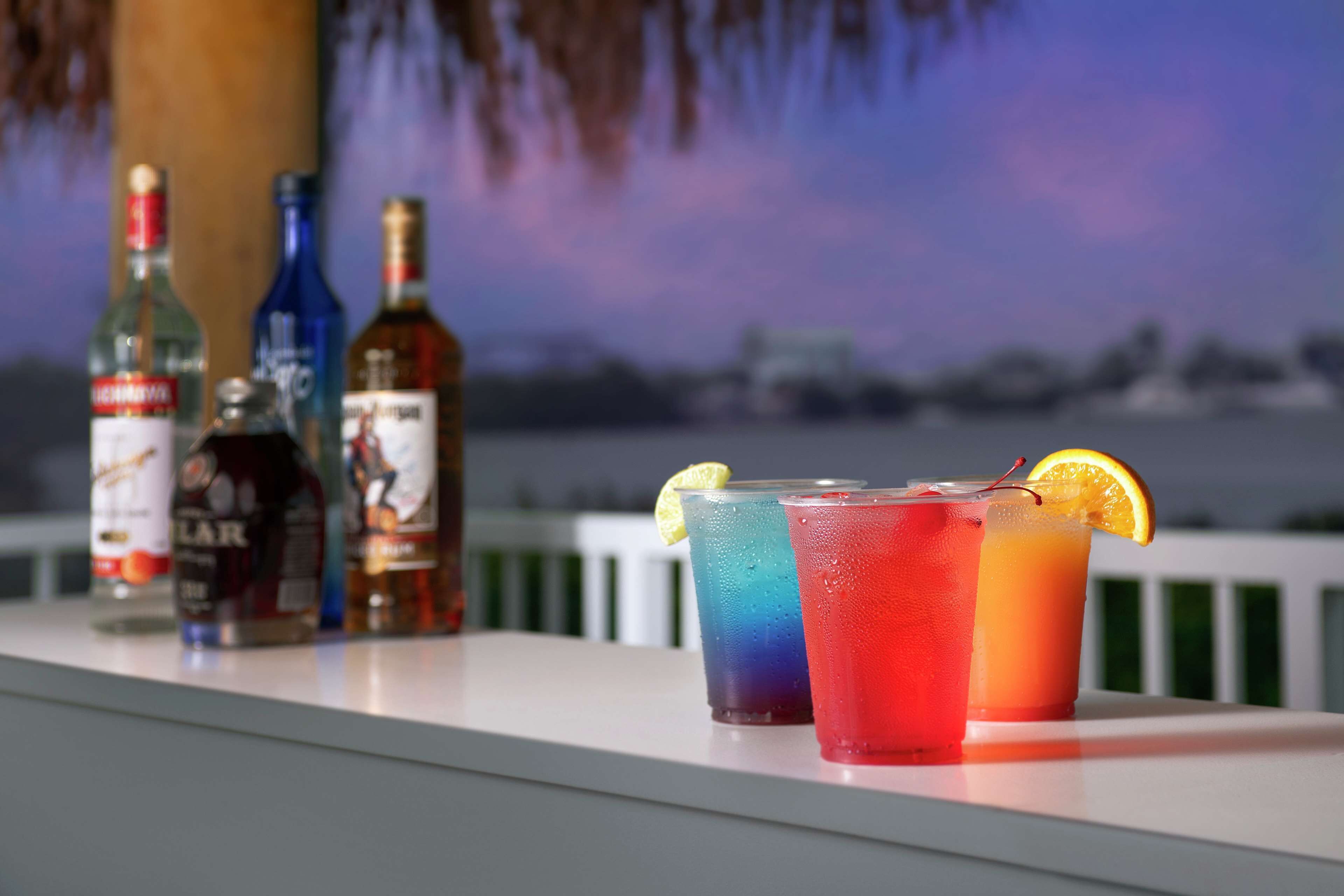 Un bar con varios botellas y bebidas coloridas sobre una barra, con vistas al cielo y edificios lejanos.