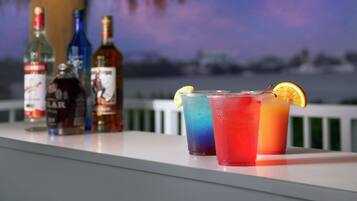 Un bar con varios botellas y bebidas coloridas sobre una barra, con vistas al cielo y edificios lejanos.
