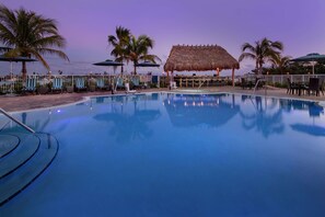 A pool area with lounge chairs and umbrellas, a thatched-roof structure, and palm trees.