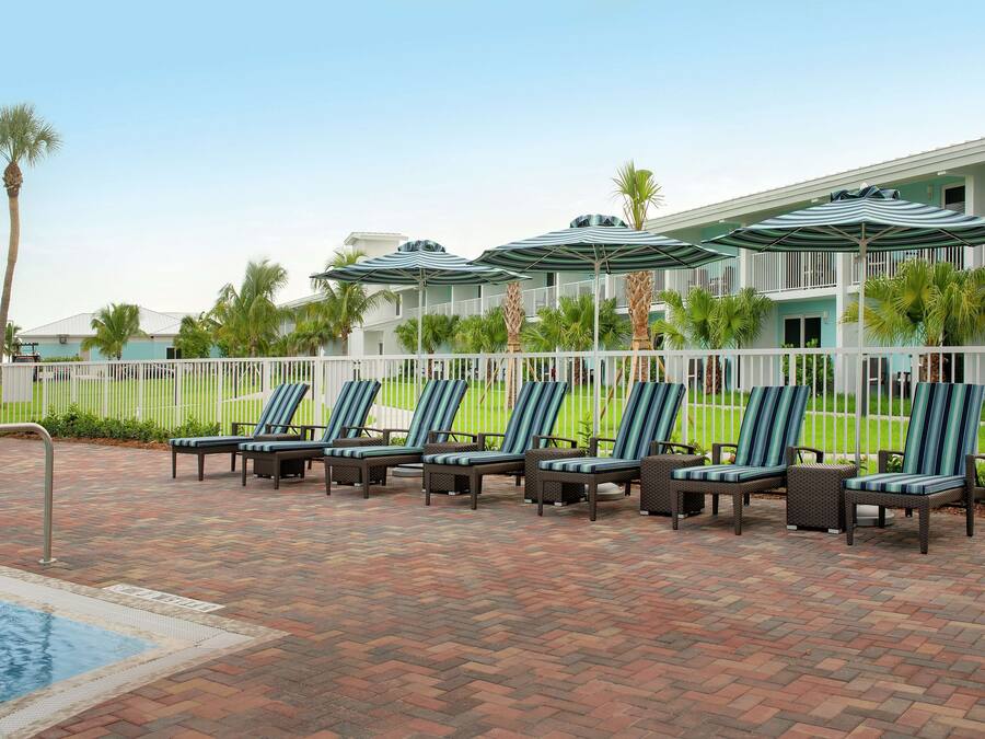 A poolside area with lounge chairs and umbrellas, a brick patio, and a building with balconies.
