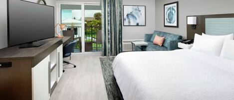 A modern hotel room with a large bed, a desk with a flat-screen TV, a sofa, and a balcony with a view of greenery.