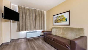 A living room with a brown leather sofa, a mounted flat-screen TV, a framed picture on the wall, and a window with patterned curtains.