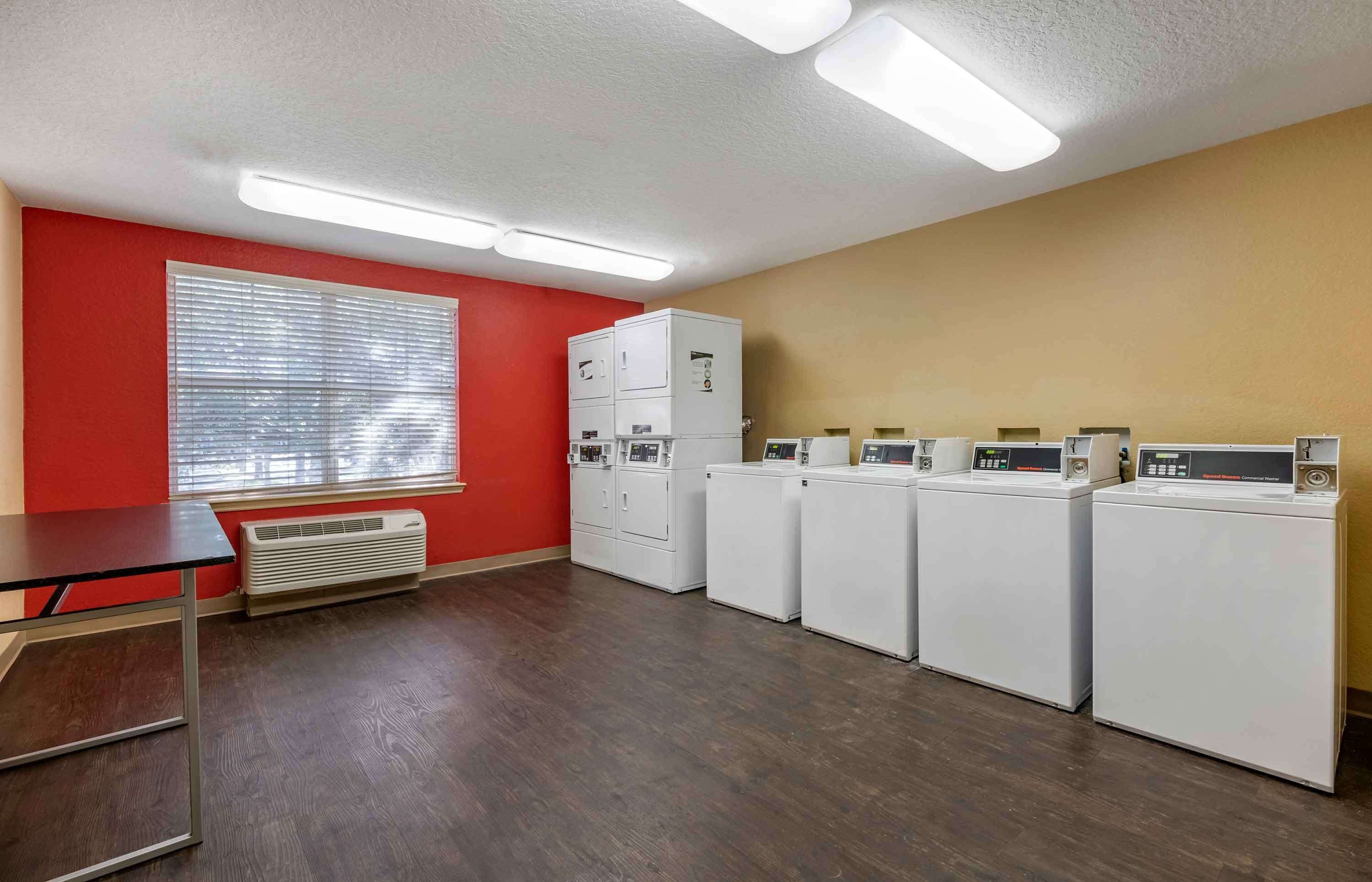 A laundry room with a table, a window with blinds, and a row of stacked washers and dryers.