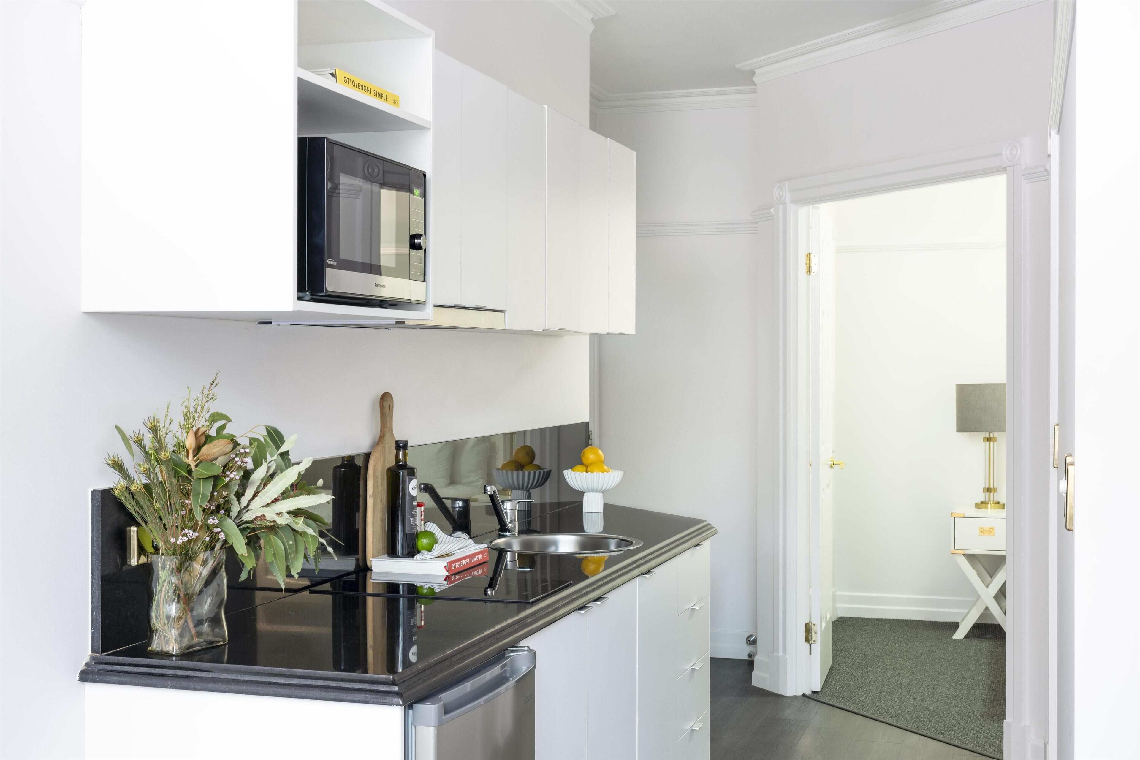 A modern kitchen with white cabinetry, a built-in microwave, and a black countertop. There is a vase with flowers on the counter, and a door leading to another room.