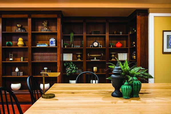 A wooden dining table with chairs, surrounded by a wooden bookshelf filled with books, decorative items, and plants.