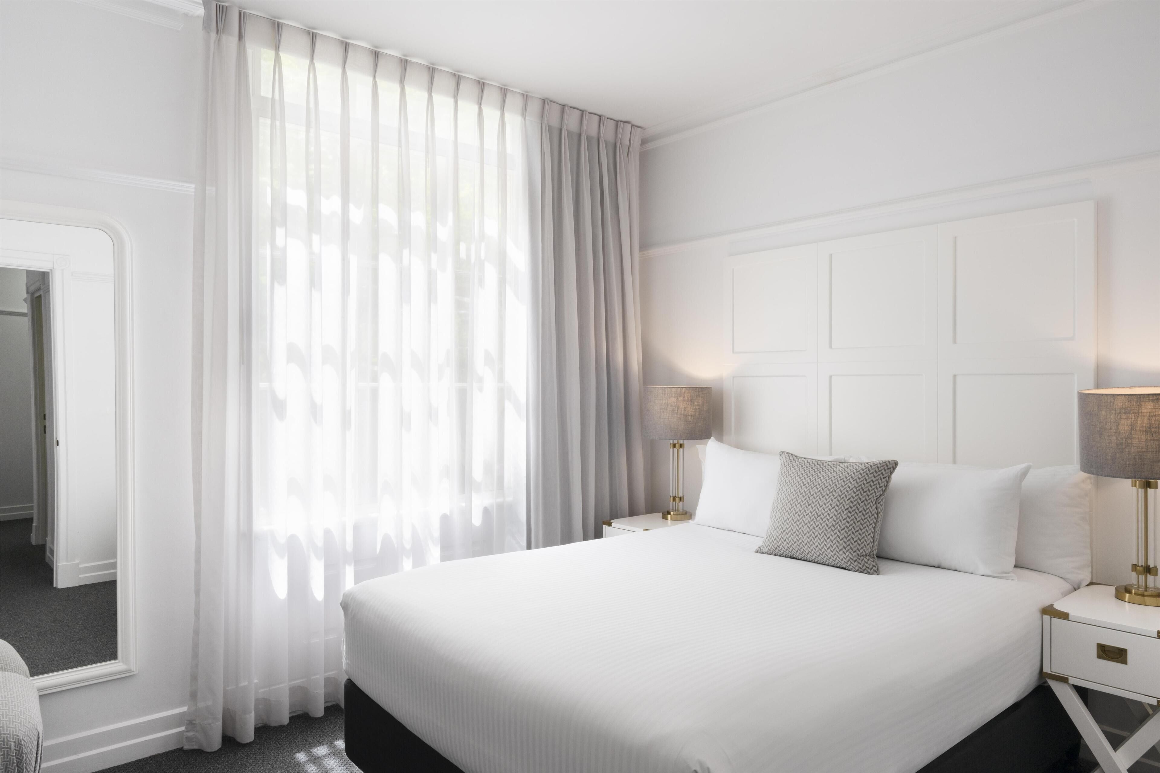 A hotel room with a large bed, white curtains, a nightstand with a lamp, and a mirror.
