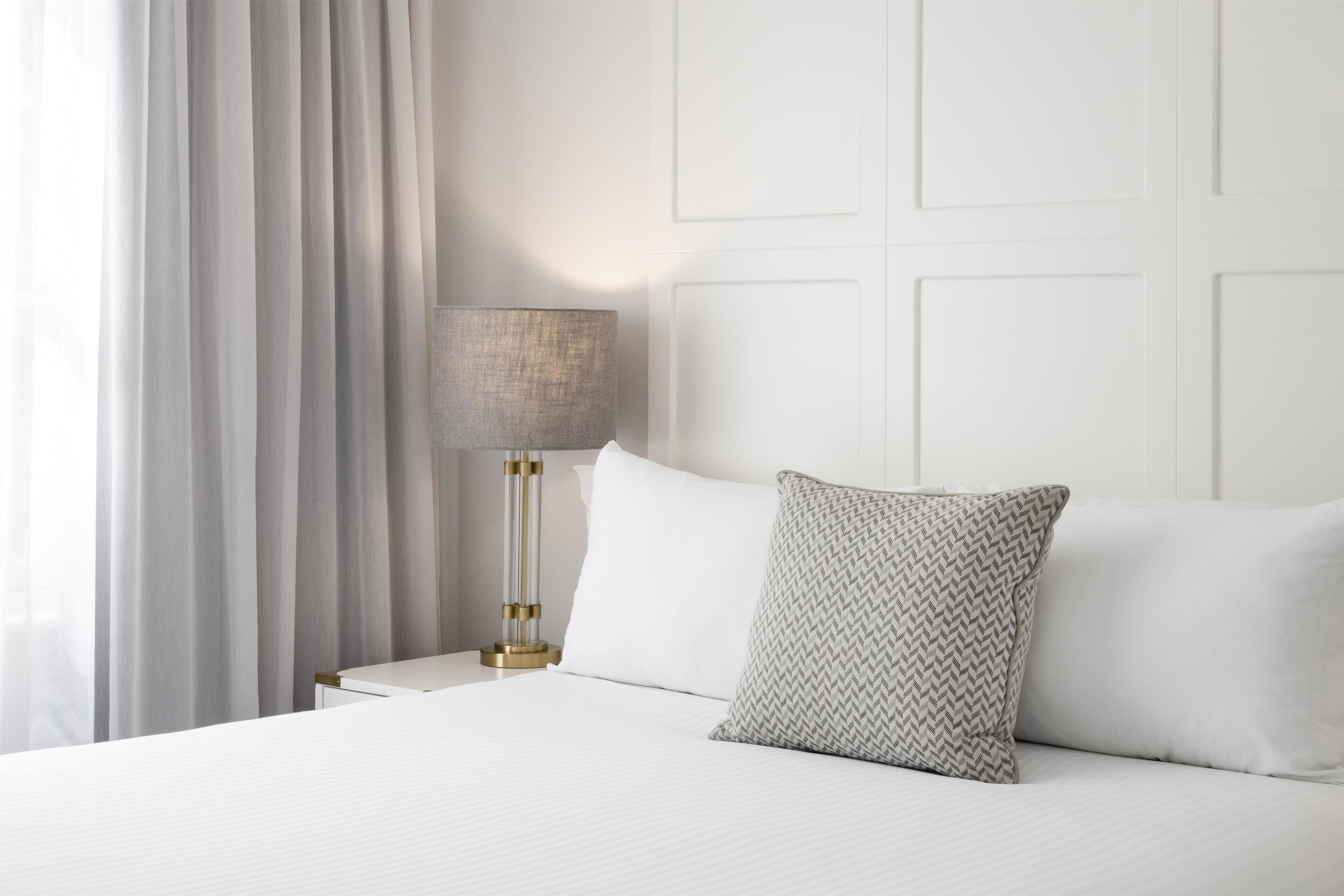 A neatly made bed with white linens and a patterned pillow, a bedside lamp with a gold base, and a white headboard.
