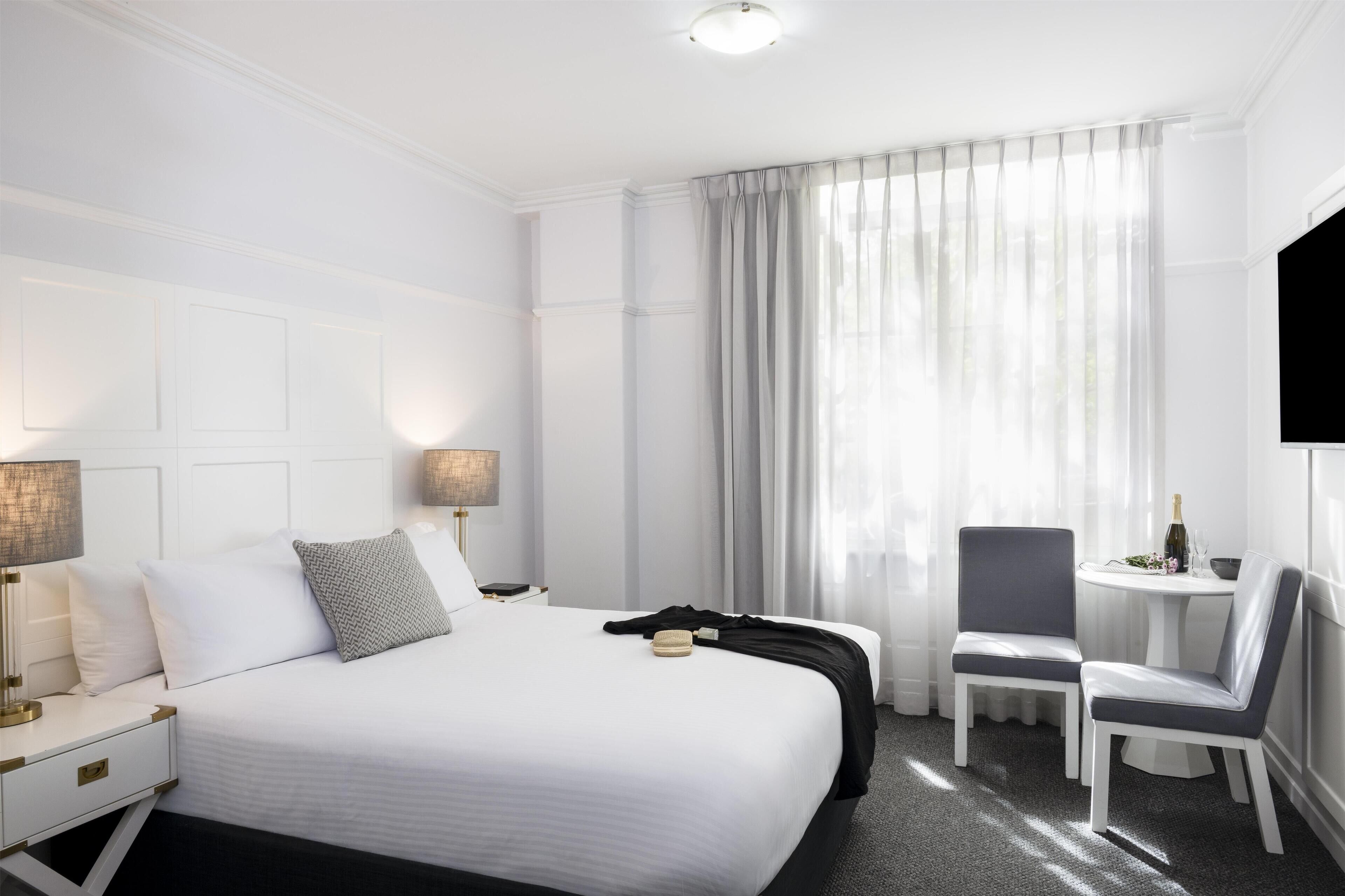 A hotel room with a large bed, bedside tables with lamps, a chair, a small table with a bottle, and a window with curtains.