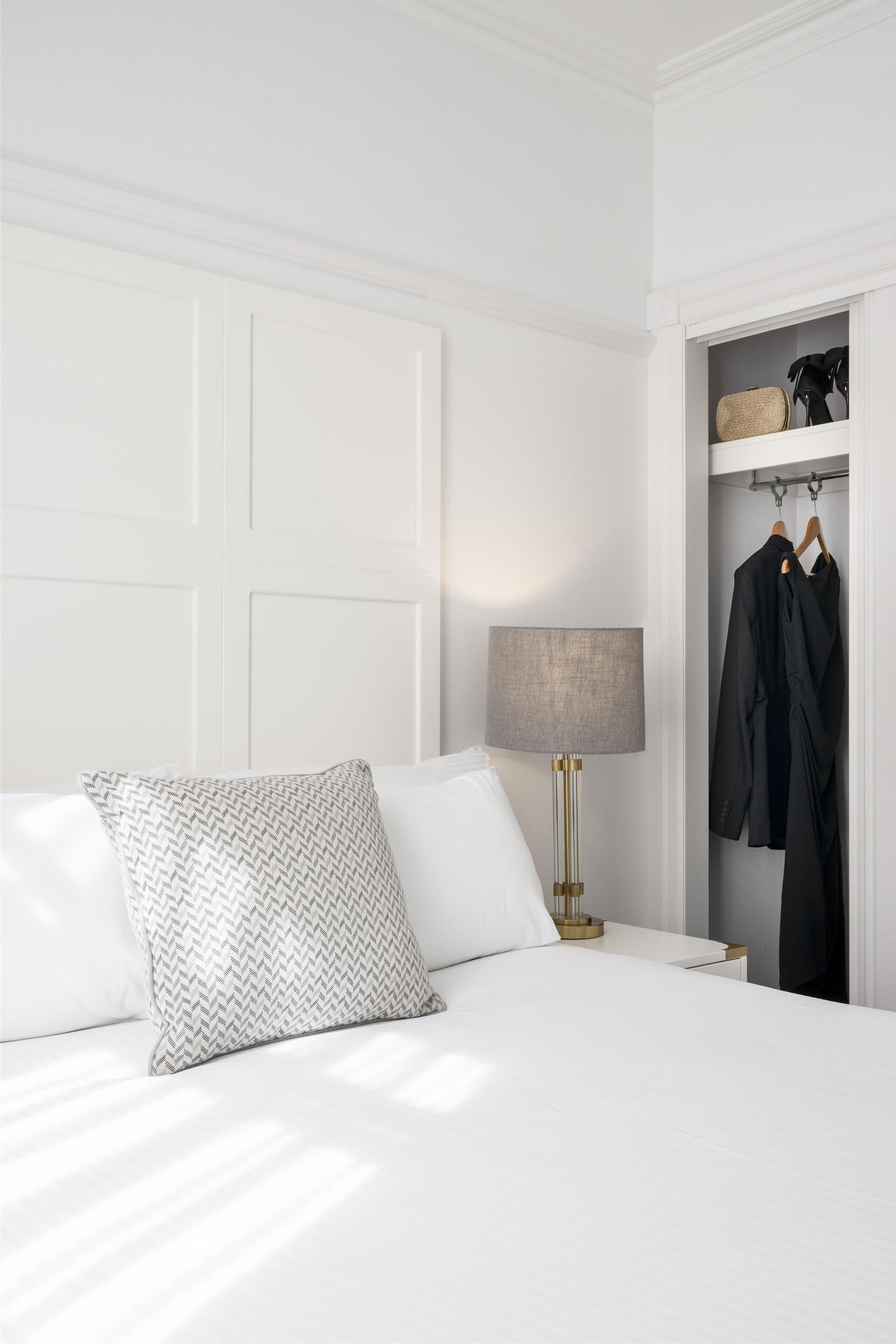 A neatly made bed with a patterned pillow, a bedside lamp, and a closet with hanging clothes.