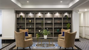 A lobby with two beige armchairs, a round table, and a shelving unit with various decorative items.