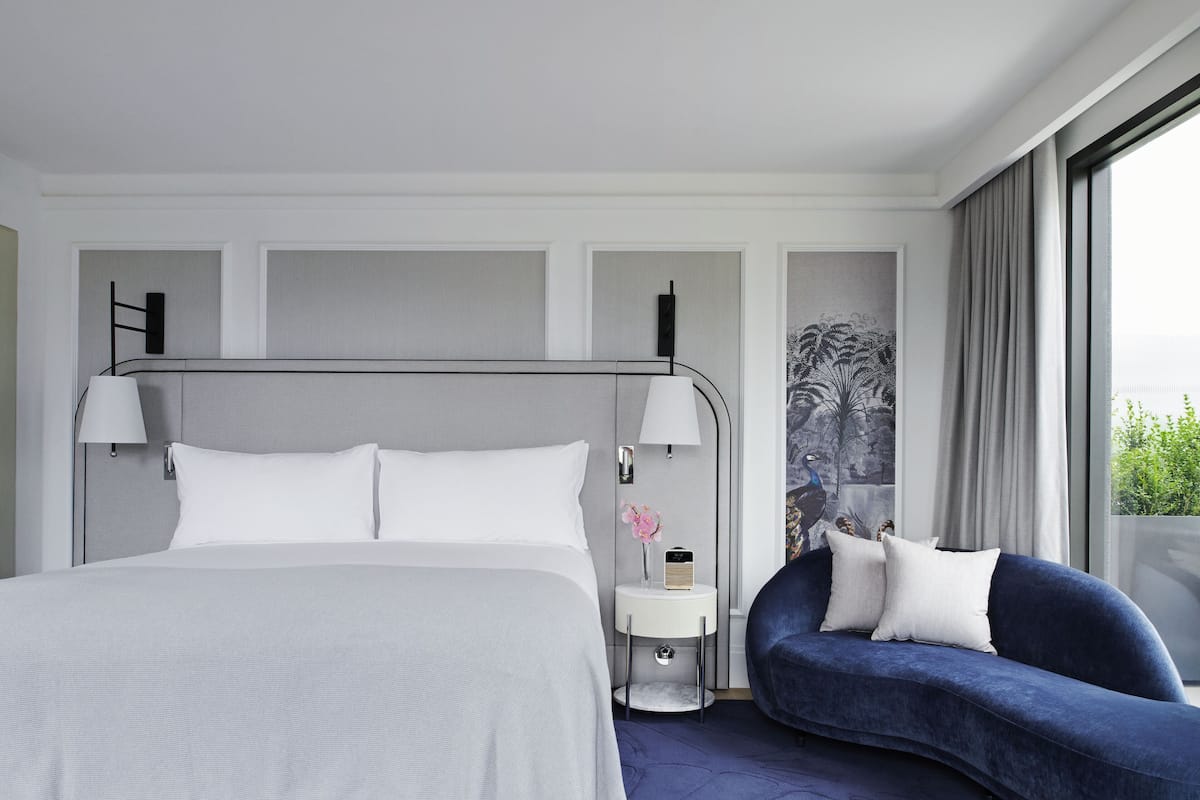 A modern hotel room with a large bed, a bedside table with a lamp, a small side table with a radio, and a blue sofa with pillows.