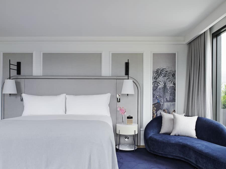 Una habitación de hotel moderna con una cama grande, una mesita de noche con lámpara, una pequeña mesa auxiliar con radio y un sofá azul con almohadones.