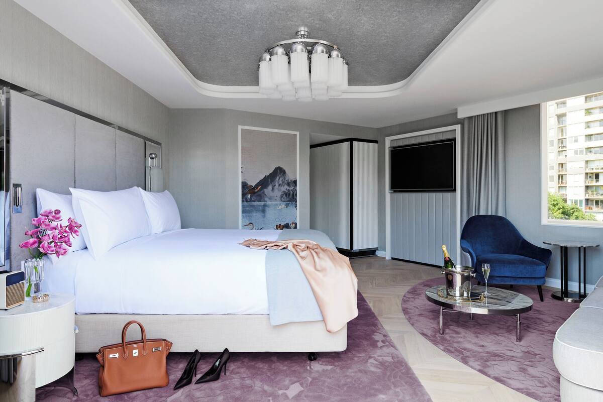 A modern hotel room with a large bed, a bedside table with a radio, a minibar, a TV, a blue armchair, and a view of the city.