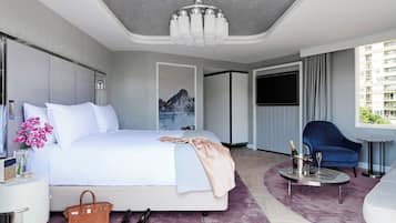 A modern hotel room with a large bed, a bedside table with a radio, a minibar, a TV, a blue armchair, and a view of the city.