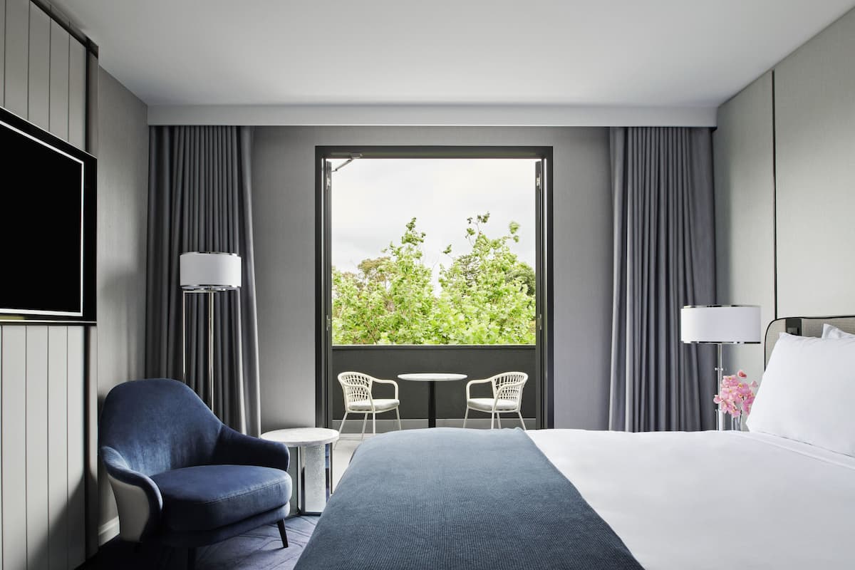 A hotel room with a large bed, a TV mounted on the wall, a small table, two chairs, a window with curtains, and a lamp.