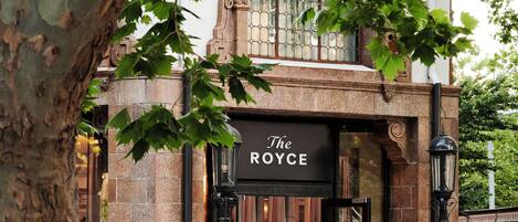 A building with a sign reading 'The Royce' at the entrance, featuring a stone facade and a large tree in the foreground.