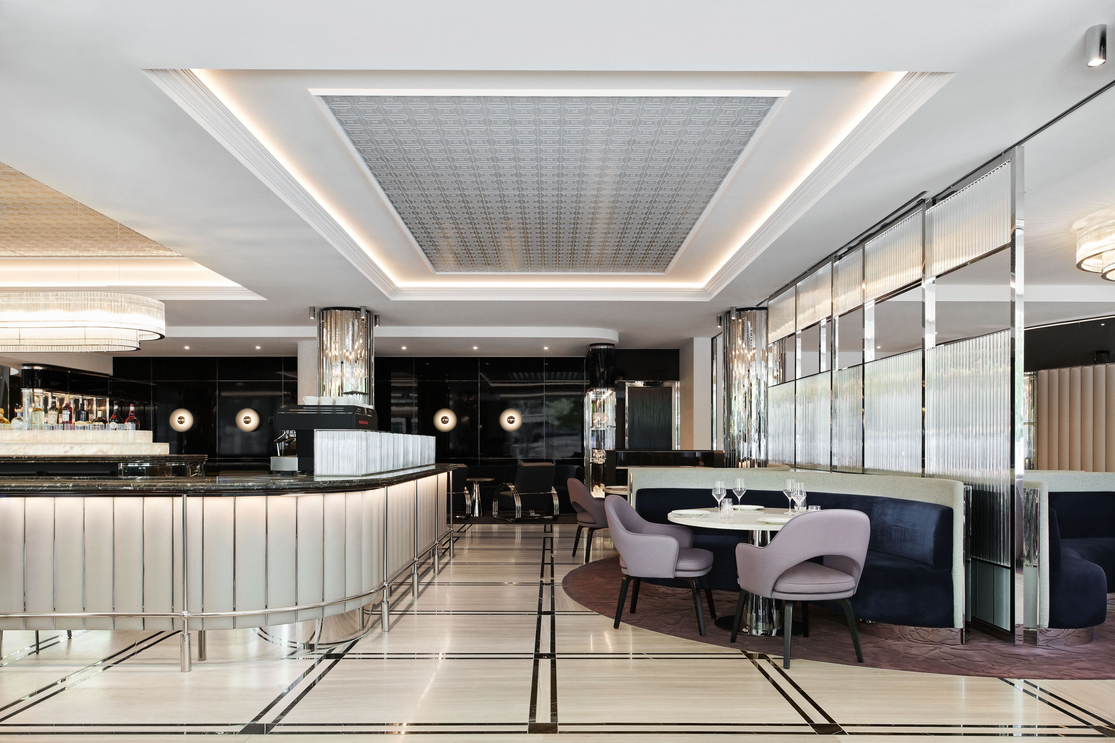 A modern bar area with a curved counter, seating with round tables, and a patterned ceiling.