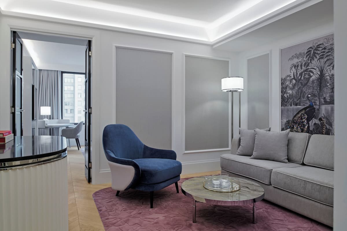 A modern hotel room with a grey sofa, a blue armchair, a glass coffee table, and a large abstract painting on the wall.