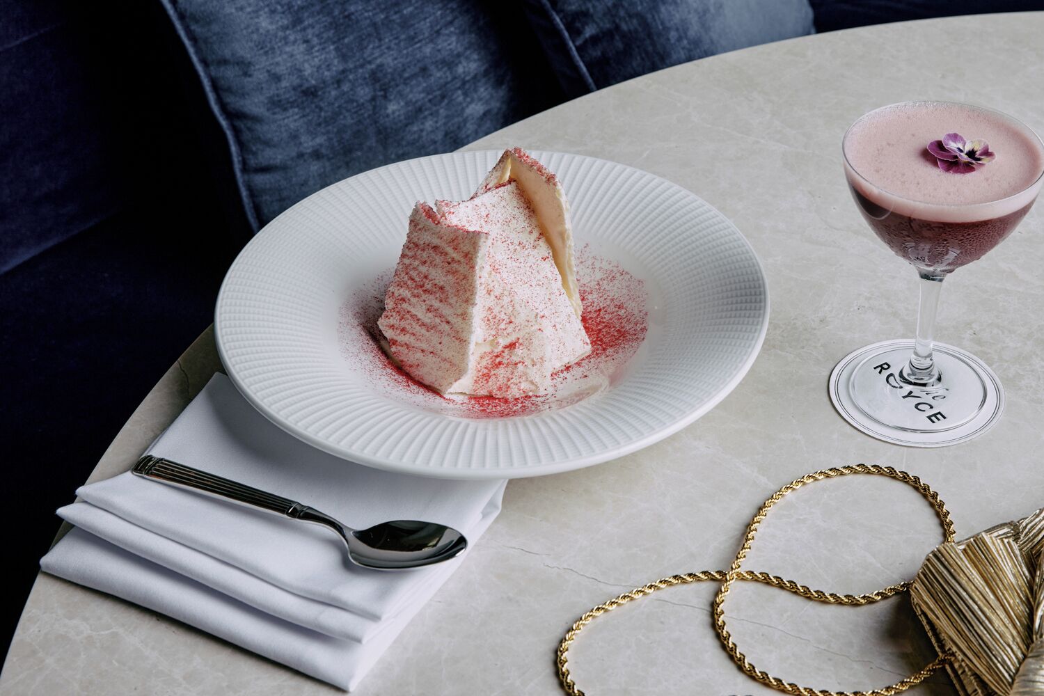 En dessert på en tallrik, ett glas med dryck, en sked, en servett och ett guldfärgat halsband på ett bord.