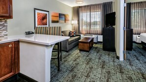 A hotel room with a kitchenette, a sofa, a coffee table, a TV, and a large window with curtains. - Staybridge Suites Lubbock - University Area by IHG (Lubbock)