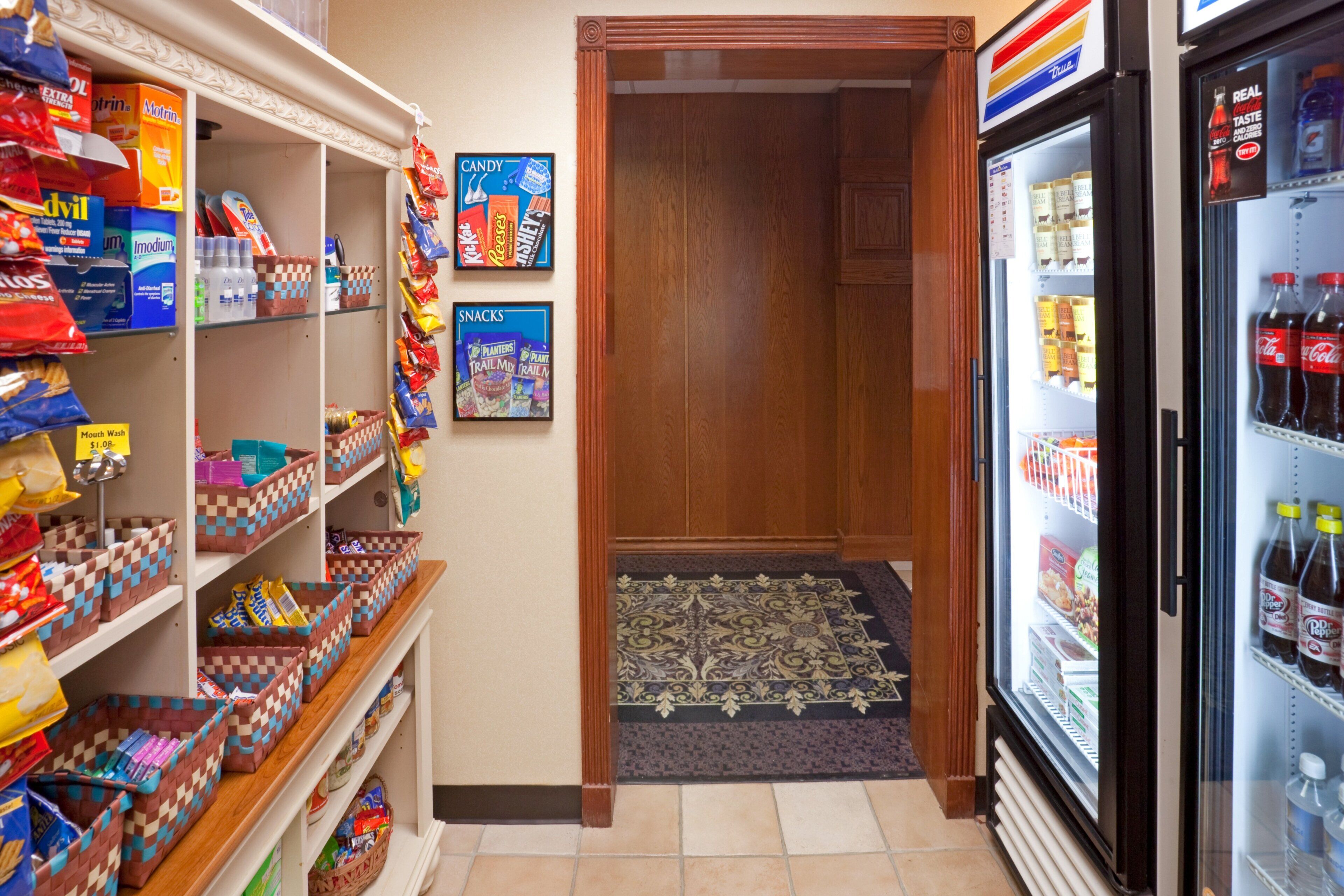A convenience store with shelves stocked with snacks, a refrigerator with beverages, and a door leading to a hallway.