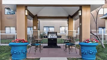 Un espace extérieur aménagé en patio, avec un barbecue, deux chaises et deux grands pots de fleurs.