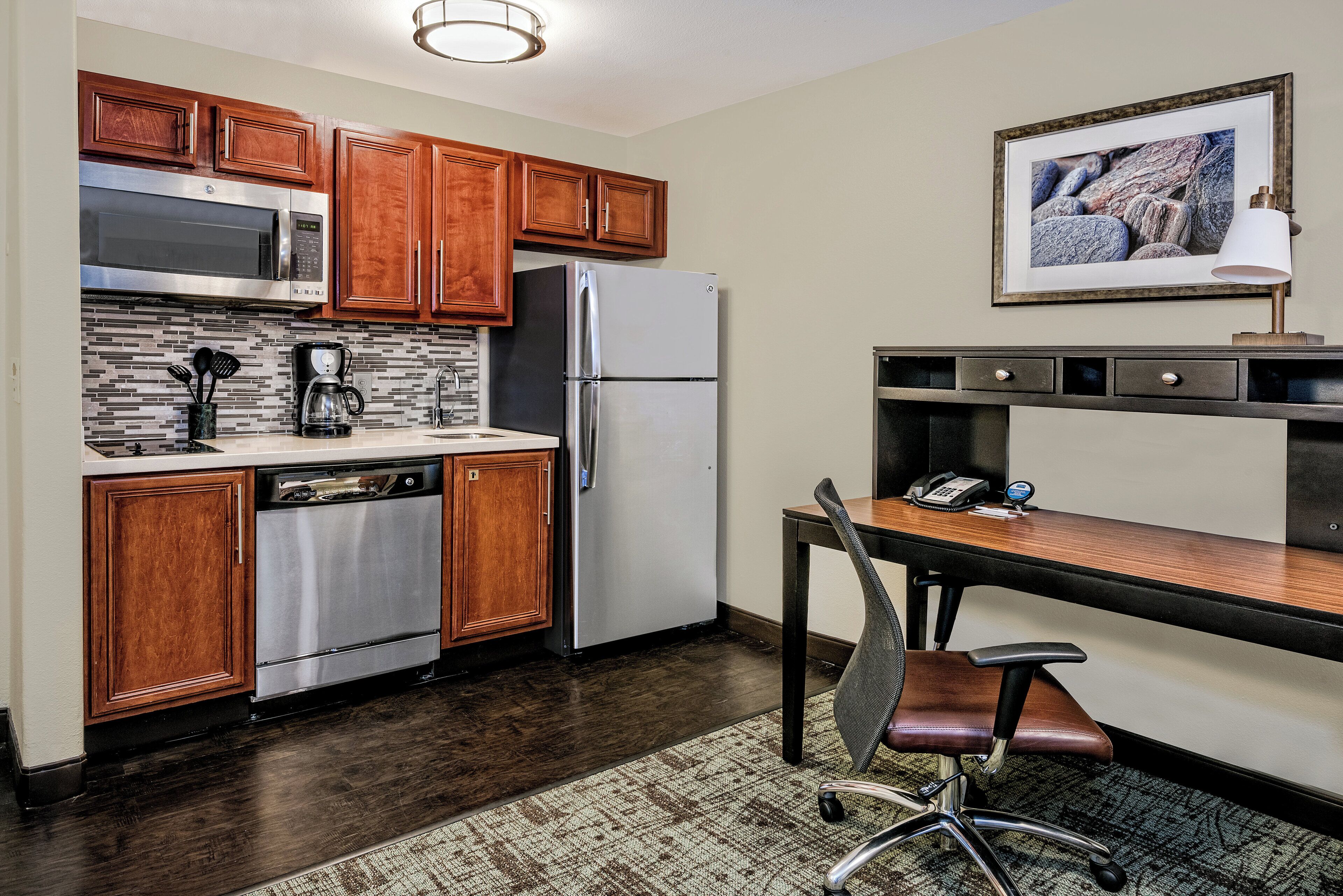 A compact kitchen with wooden cabinets, a stainless steel refrigerator, a coffee maker, and a small dining area with a chair and table.