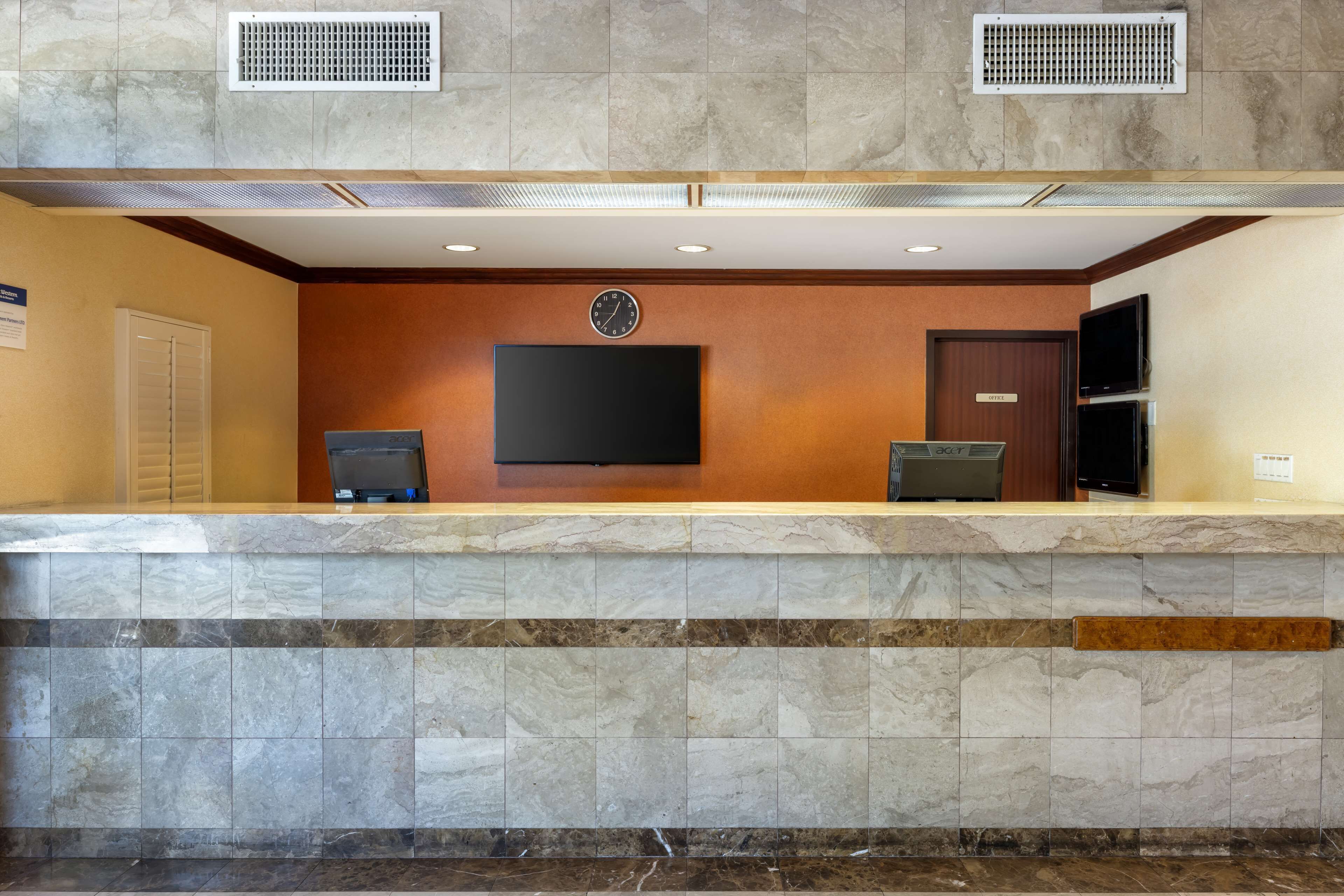 Zona de recepción de un hotel con mostrador de mármol, televisor de pantalla plana, un reloj y una puerta con la etiqueta 'EXIT'.