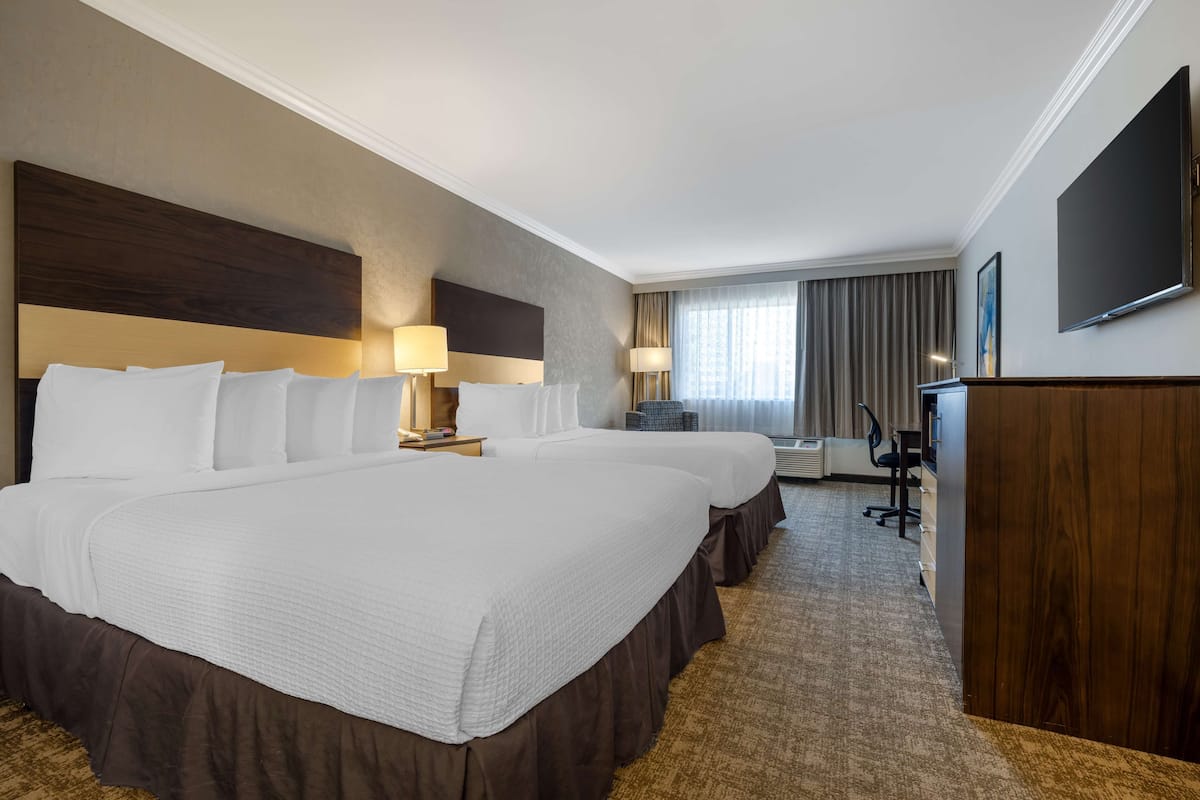 A hotel room with a large bed, two bedside tables with lamps, a desk with a chair, a flat-screen TV, and a window with curtains.