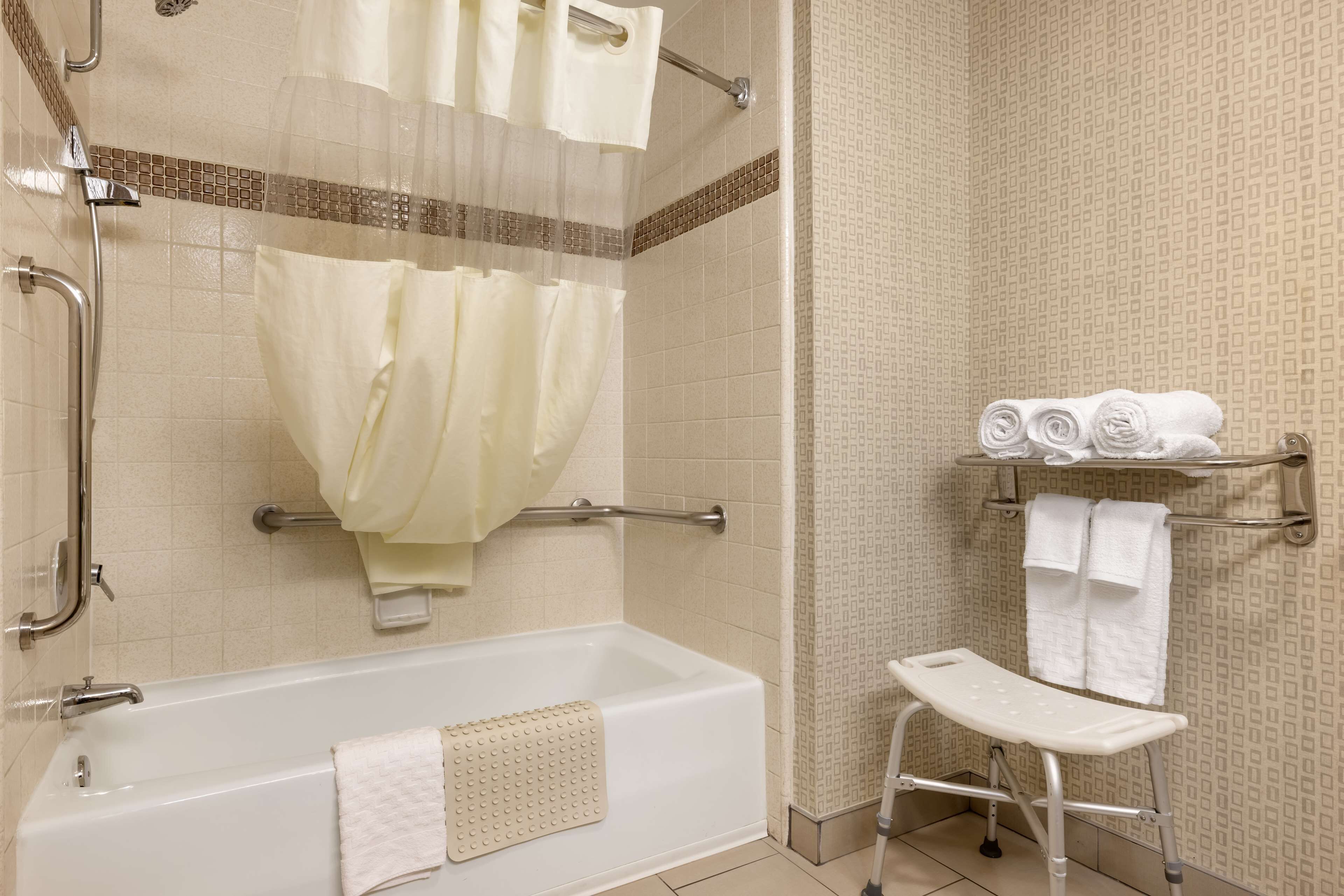 Une salle de bain équipée d’une baignoire, d’une douche avec une porte en verre, d’une barre d’appui, d’une étagère avec des serviettes et d’un banc accessible aux personnes en fauteuil roulant.