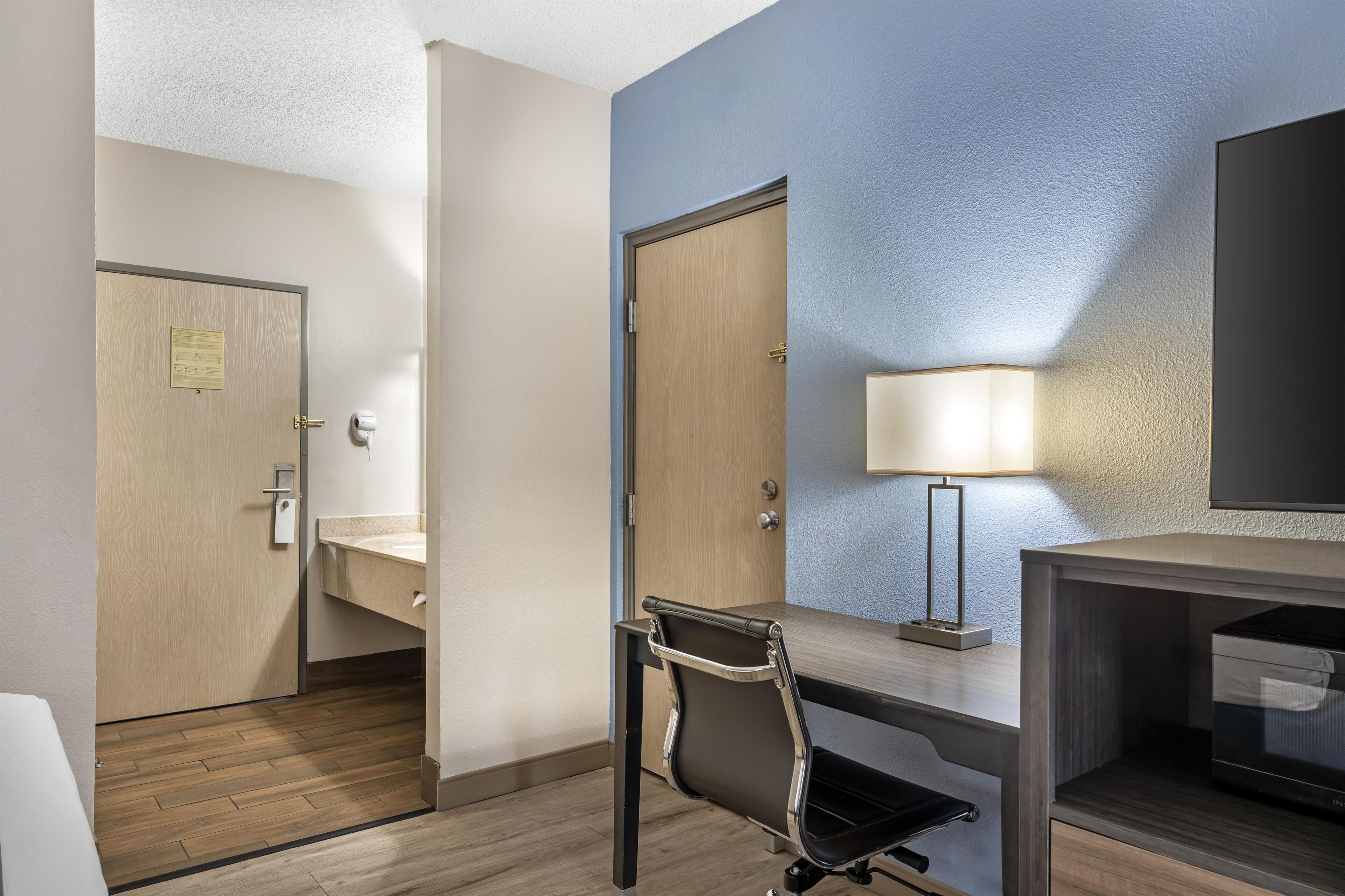 A hotel room with a desk, chair, bedside table, lamp, and a television.