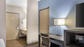 A hotel room with a desk, chair, bedside table, lamp, and a television.