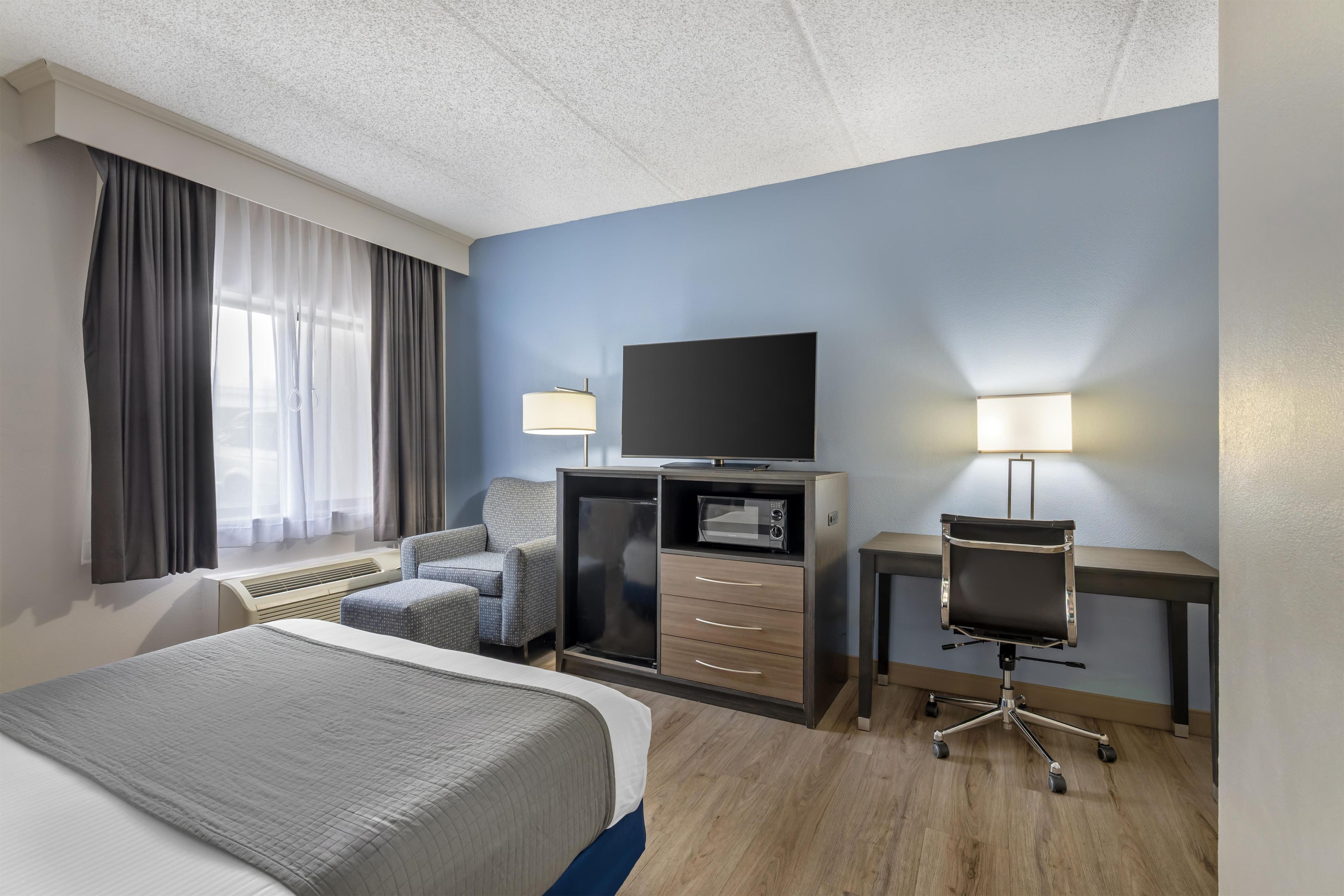 A hotel room with a bed, a desk with a flat-screen TV, a chair, a microwave, and a desk lamp.
