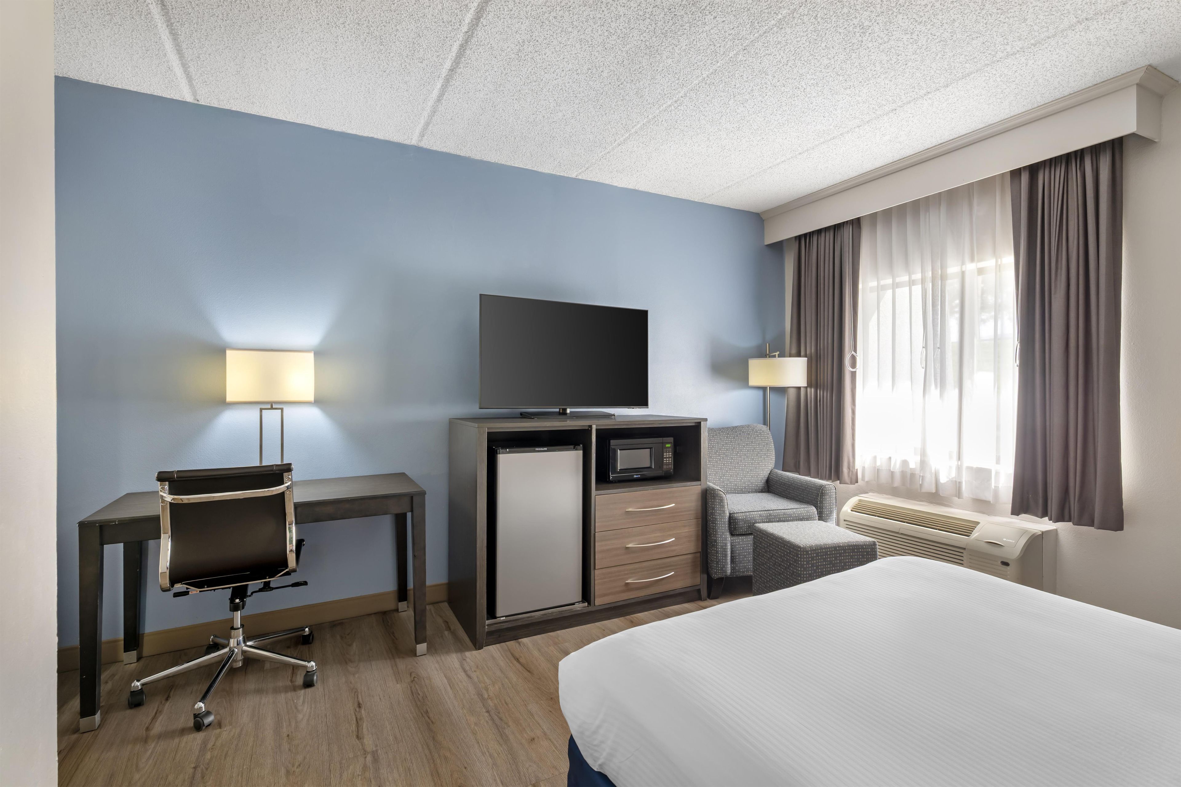 A hotel room with a bed, a desk with a chair, a television, a mini-fridge, a lamp, and a window with curtains.
