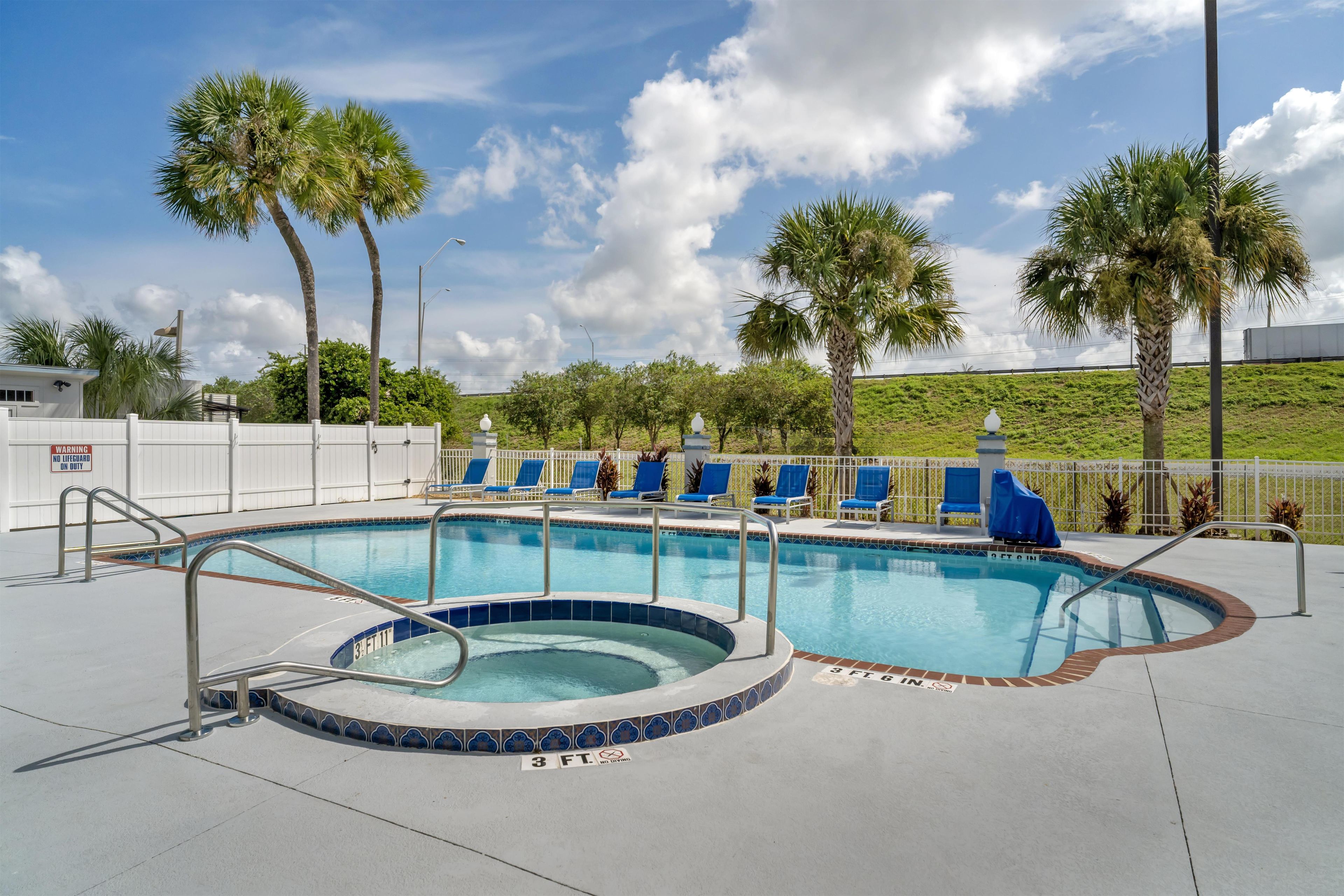 A swimming pool with a hot tub, surrounded by lounge chairs, palm trees, and a white fence.