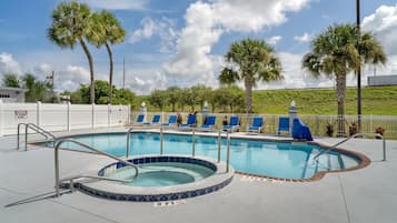 A swimming pool with a hot tub, surrounded by lounge chairs, palm trees, and a white fence.