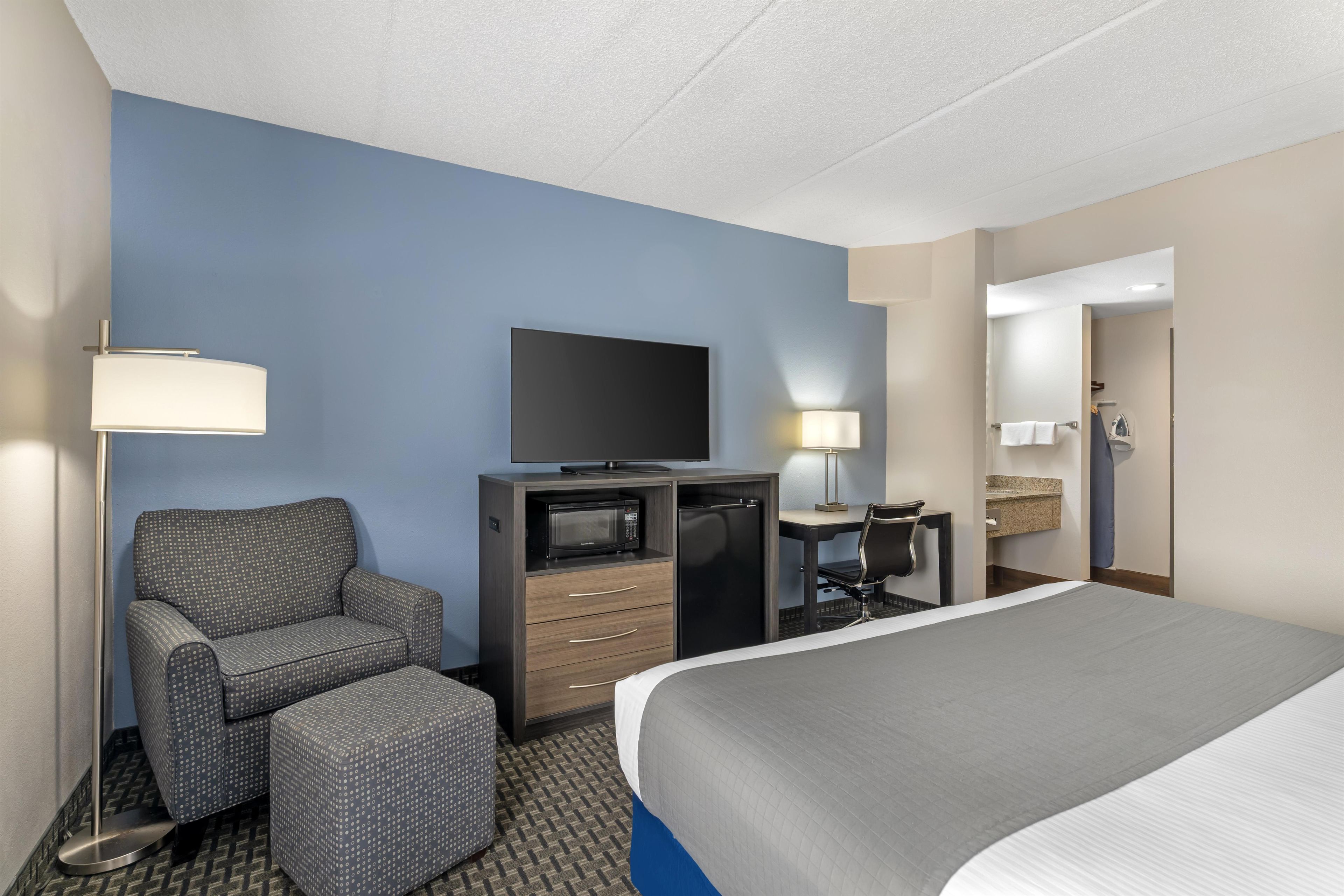 A hotel room with a bed, a desk with a flat-screen TV, a chair, a microwave, and a lamp.