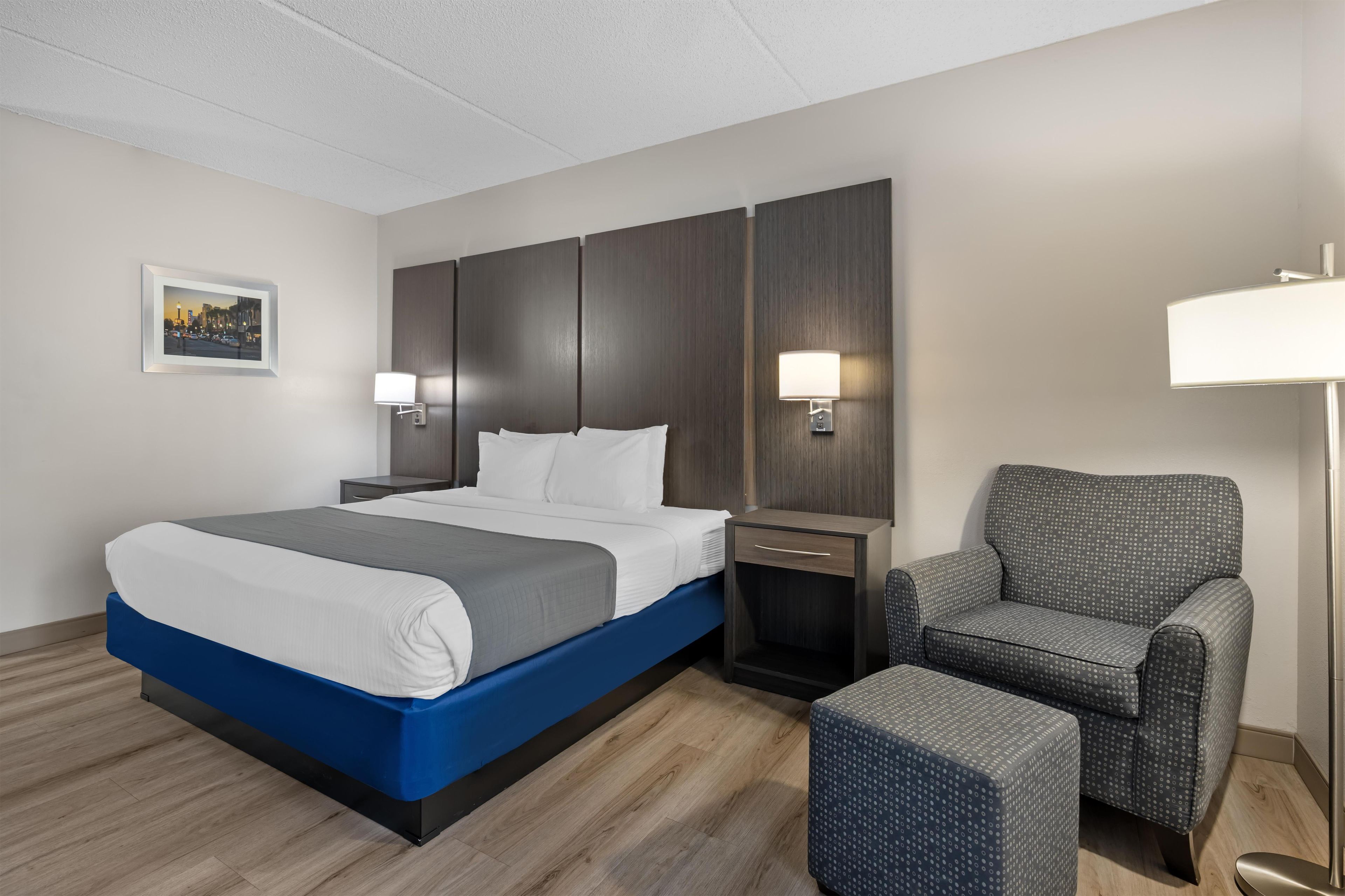 A hotel room with a bed, a chair, a nightstand, a lamp, and a picture on the wall.