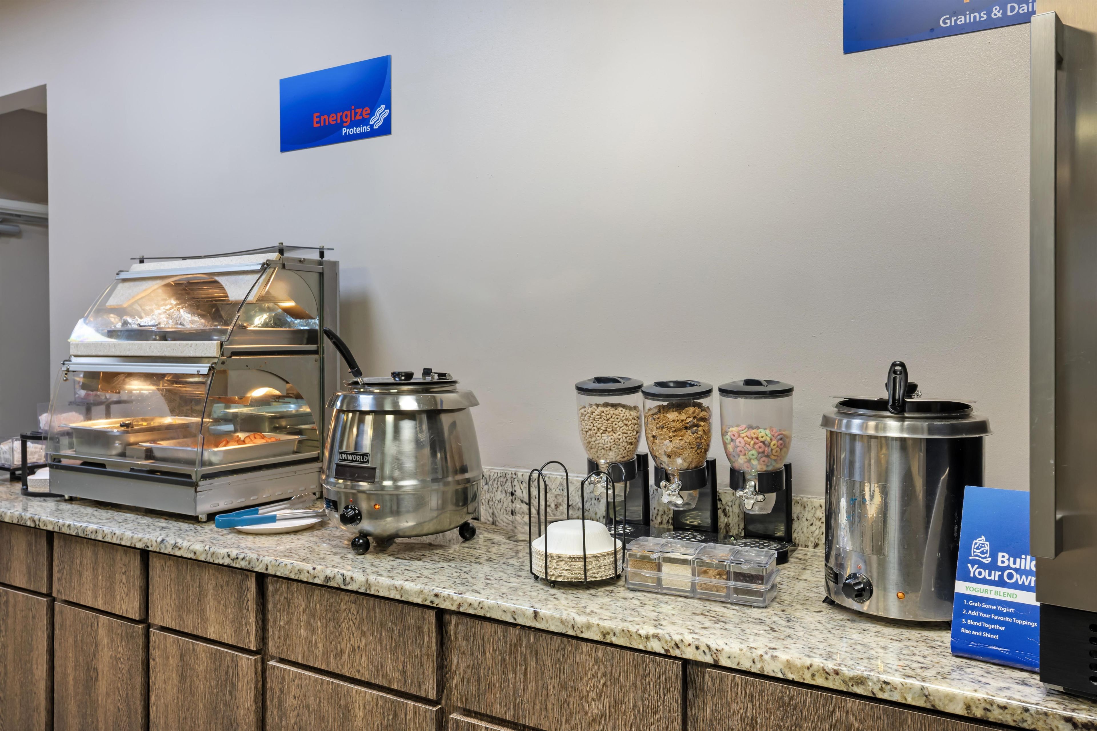A breakfast buffet setup with a self-service yogurt station, cereal dispensers, and a hot food station.