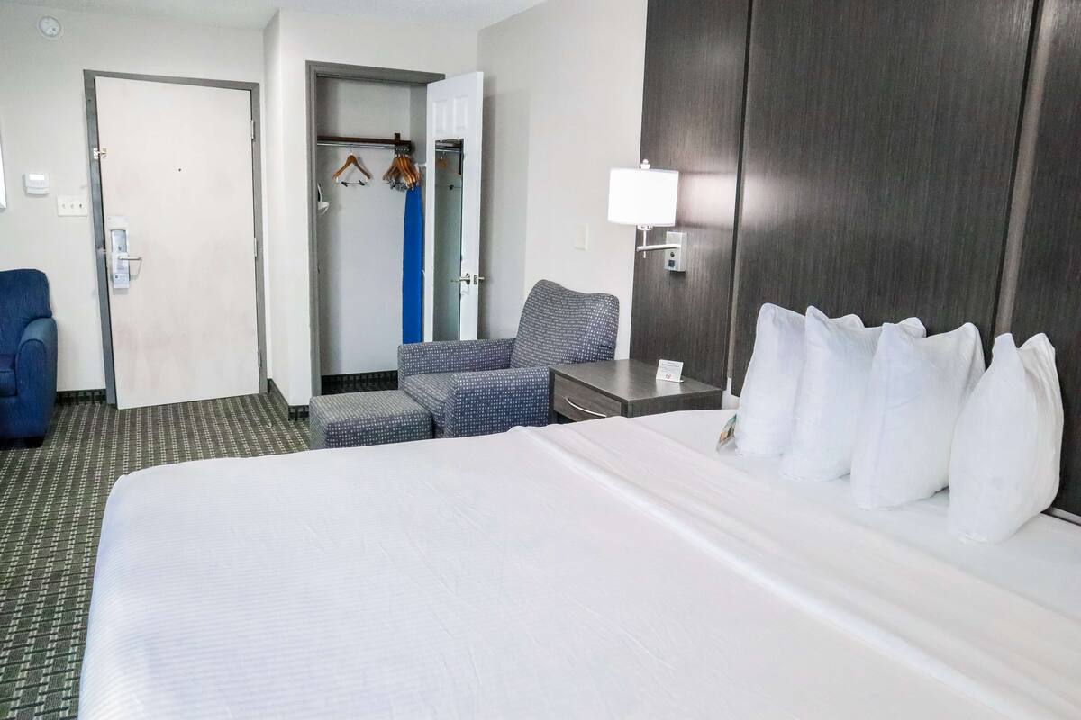 Quarto de hotel com uma cama grande, uma mesa com luminária, uma cadeira, um guarda-roupa e um banheiro visível por uma porta aberta.