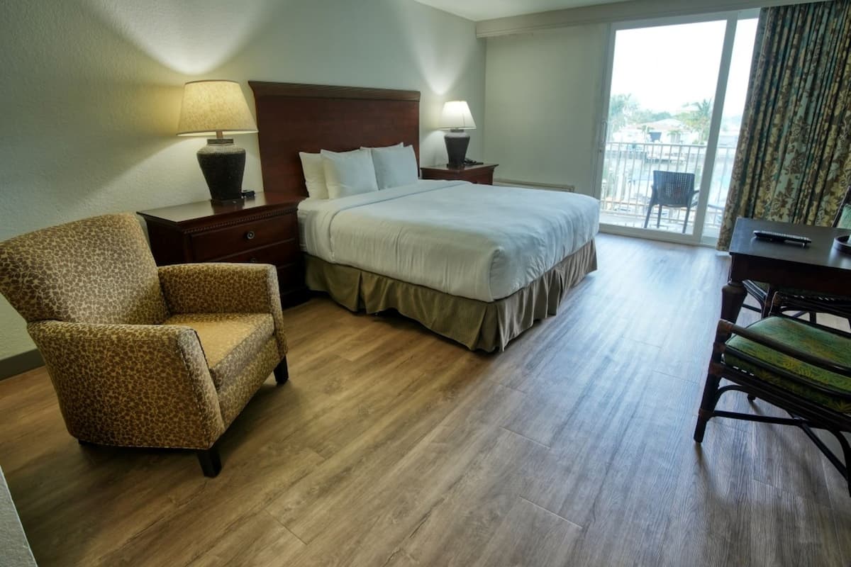 Habitación de hotel con cama, dos mesitas de noche, una silla, un escritorio y vistas al exterior a través de un ventanal grande.