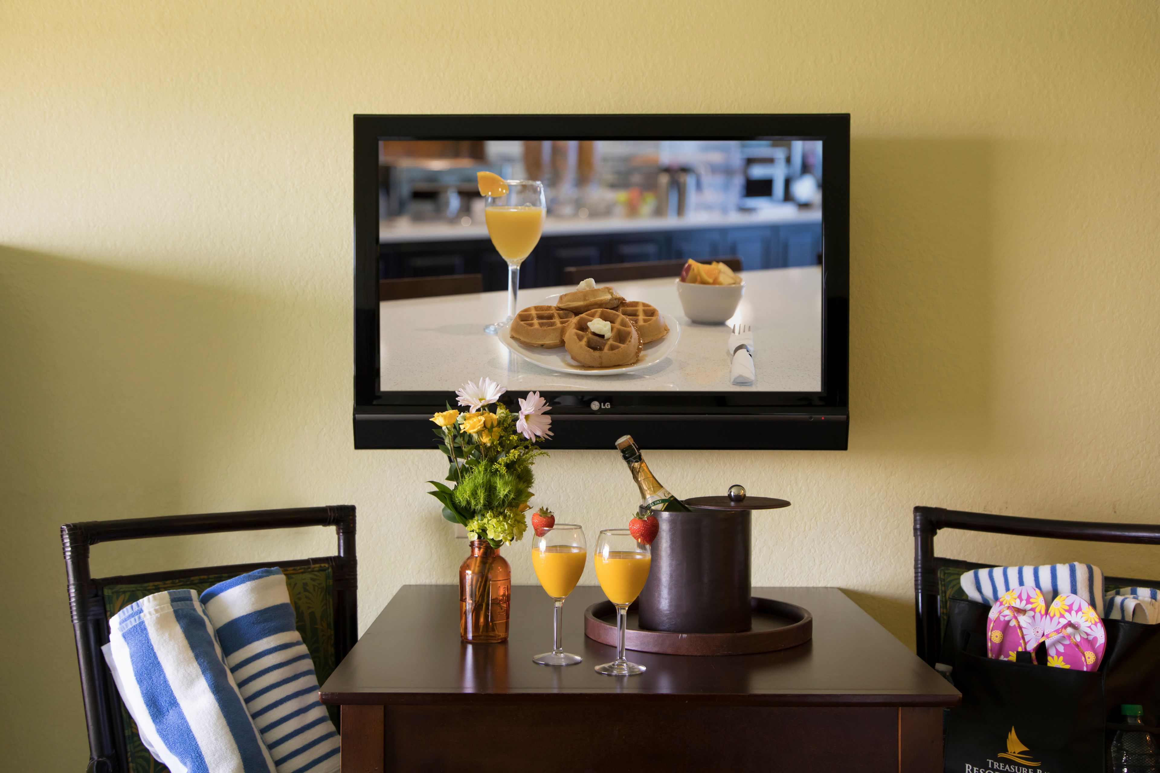 A breakfast setup with waffles, orange juice, and champagne on a tray, with a TV displaying a similar scene in the background.