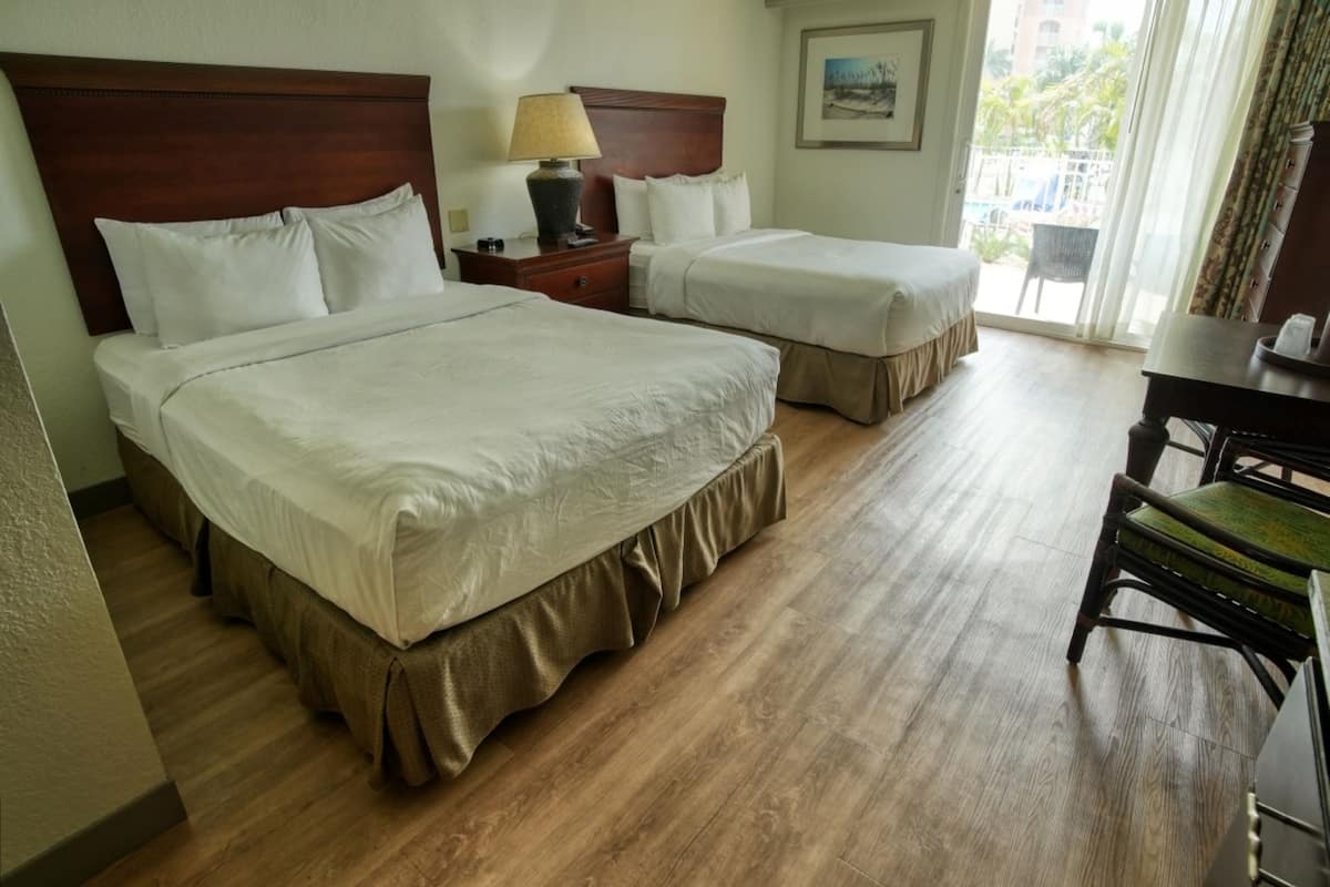 Habitación de hotel con dos camas, un escritorio, una silla y ventana con vista a palmeras.