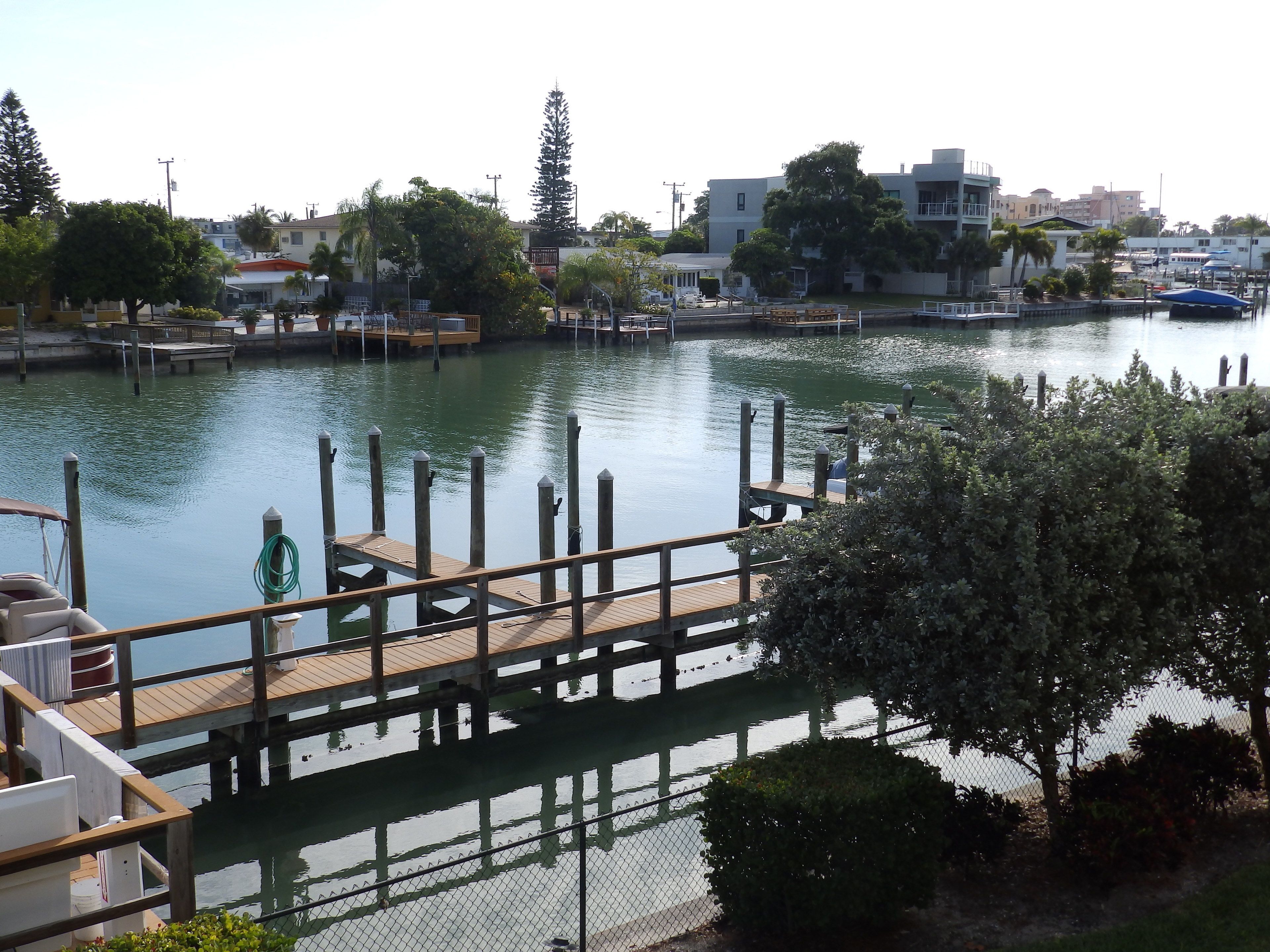 Eine Uferpromenade mit Holzsteg, einem Zaun aus Maschendraht und Blick auf Wasser und Gebäude im Hintergrund.