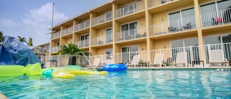Zona de piscina de hotel con agua azul cristalina, sillas de descanso y un edificio de varios pisos con balcones.