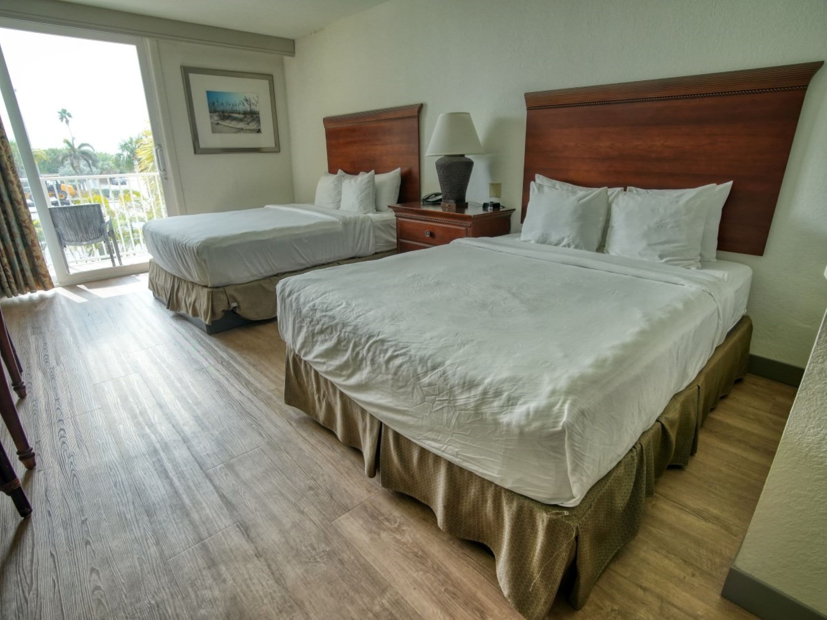 Une chambre d’hôtel avec deux lits, une tête de lit en bois, une table de chevet avec une lampe et un balcon offrant une vue sur les palmiers.