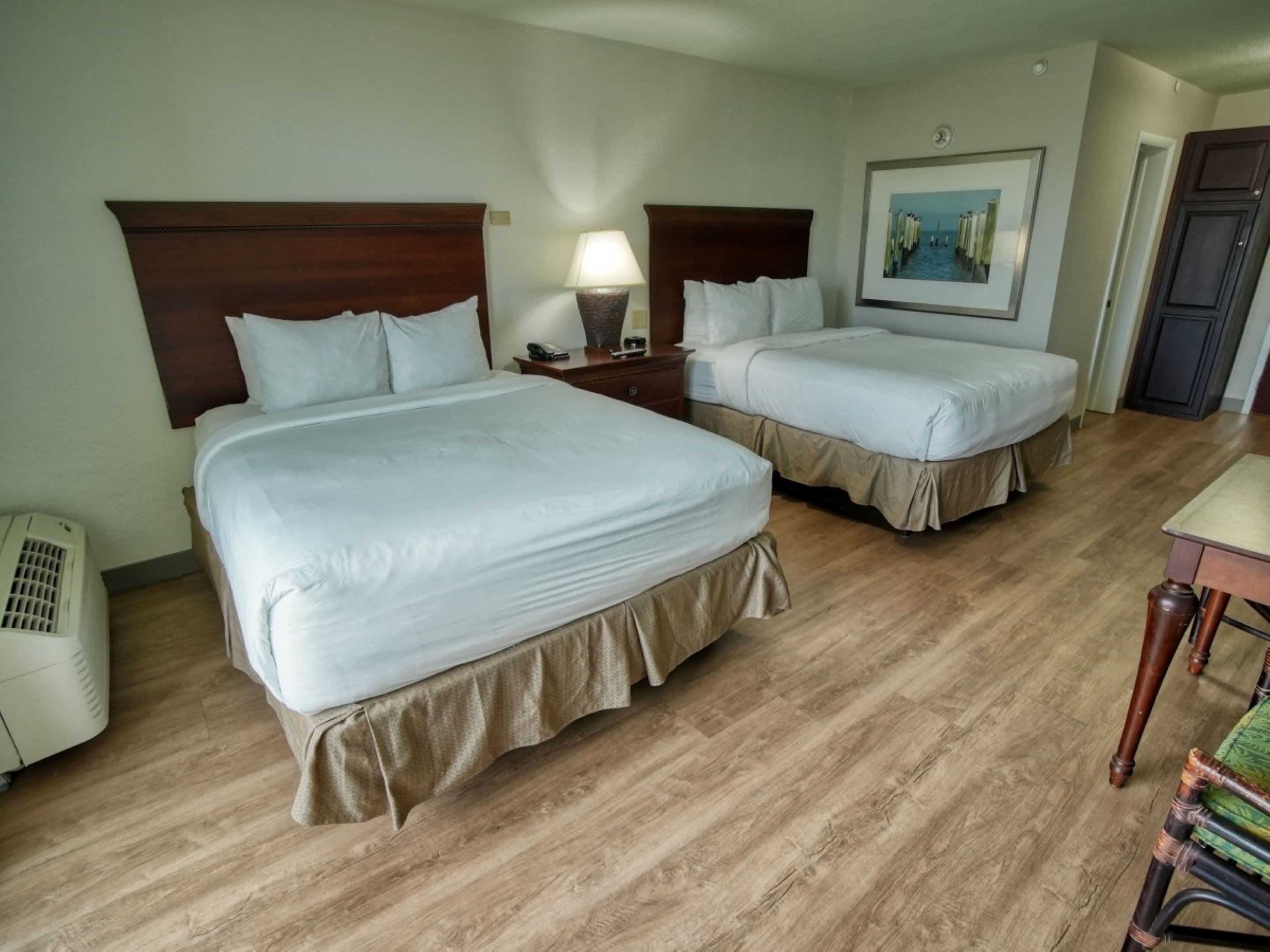 Una habitación de hotel con dos camas, una mesita de noche con lámpara, un cuadro en la pared y suelo de madera.